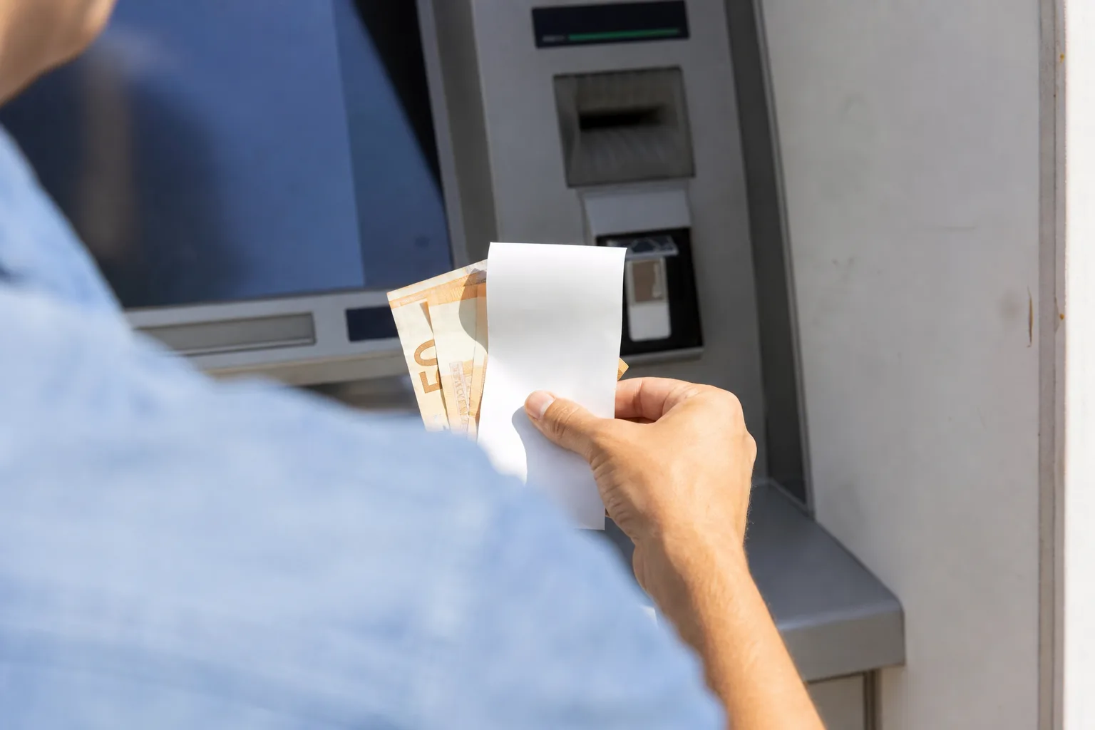 Over-the-shoulder view of someone collecting cash and a blank receipt from an ATM in bright daylight.