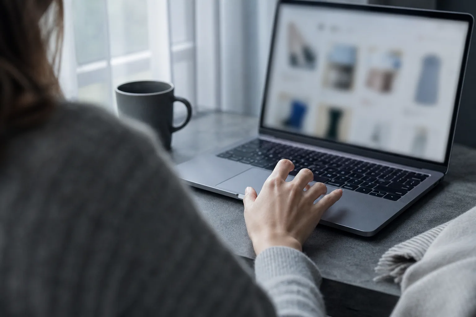 Over-the-shoulder view of a hand pausing above a laptop during an online shopping moment in cool morning light.