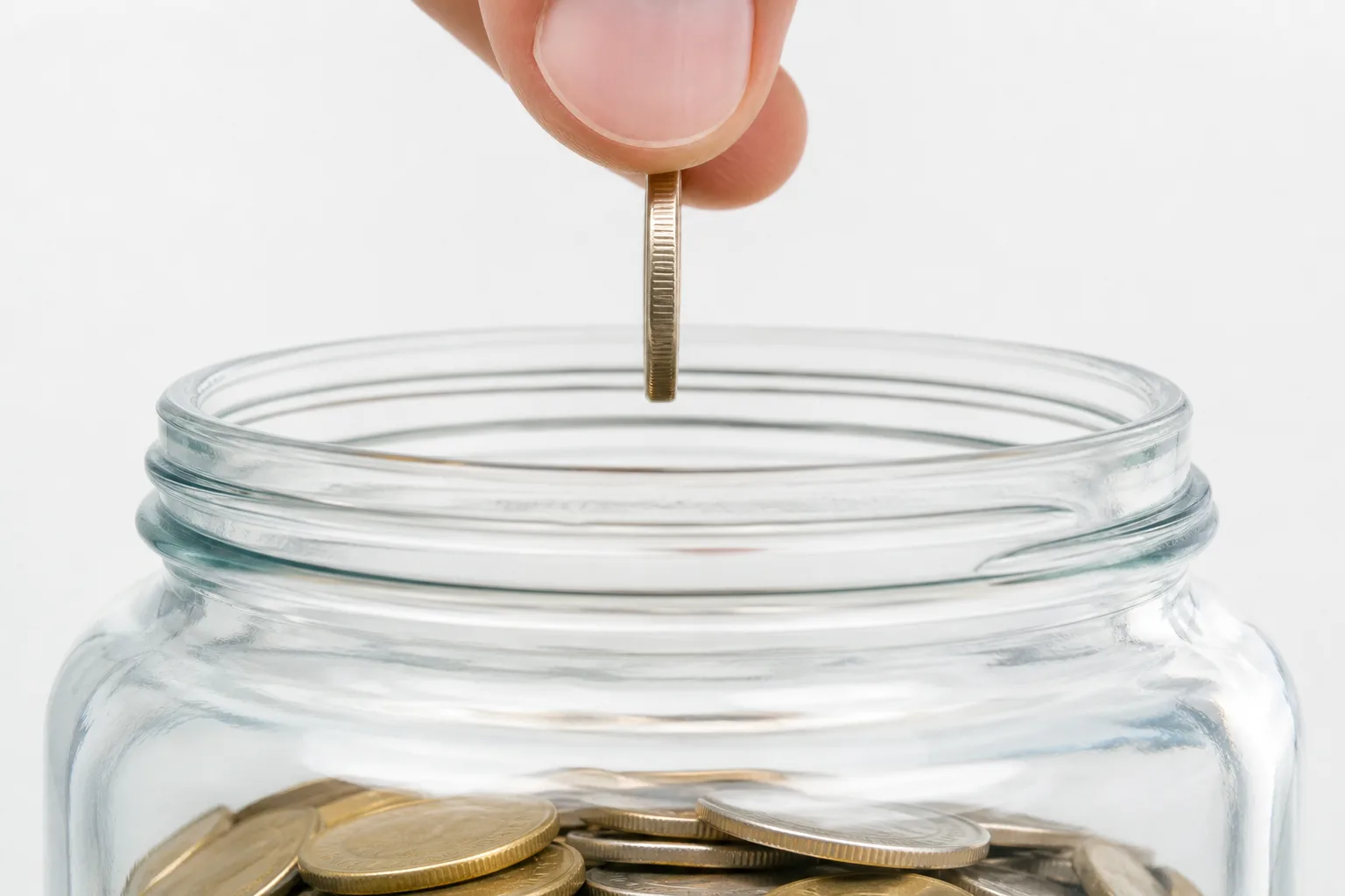 A clear savings jar fills the frame as an anonymous hand adds one coin in bright daylight.