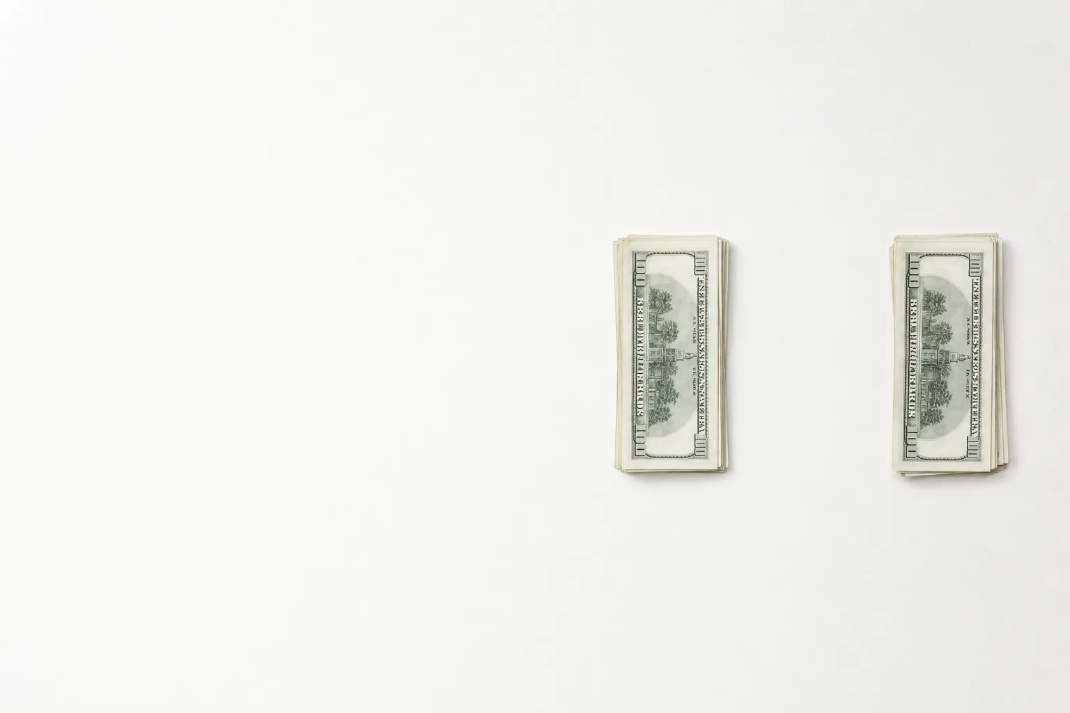 Two separated piles of unmarked cash sit on the right side of a bright white desk with an empty gap between them.
