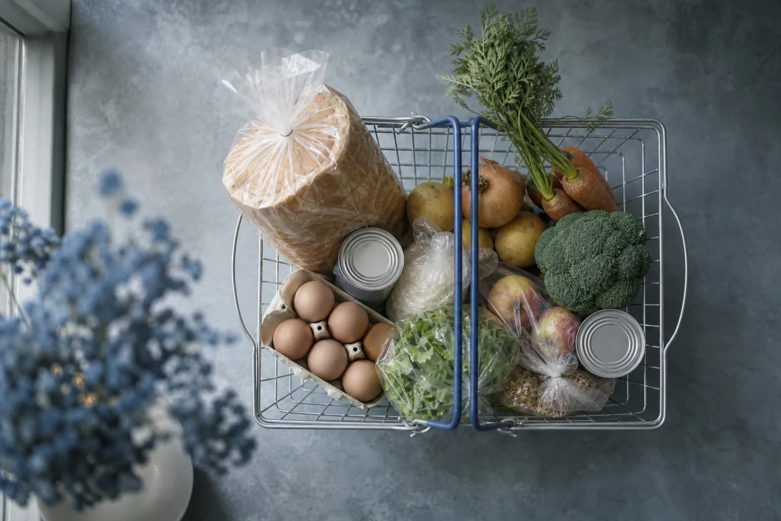 A small basket of basic groceries rests on a kitchen table in cool morning light, emphasizing everyday essentials.